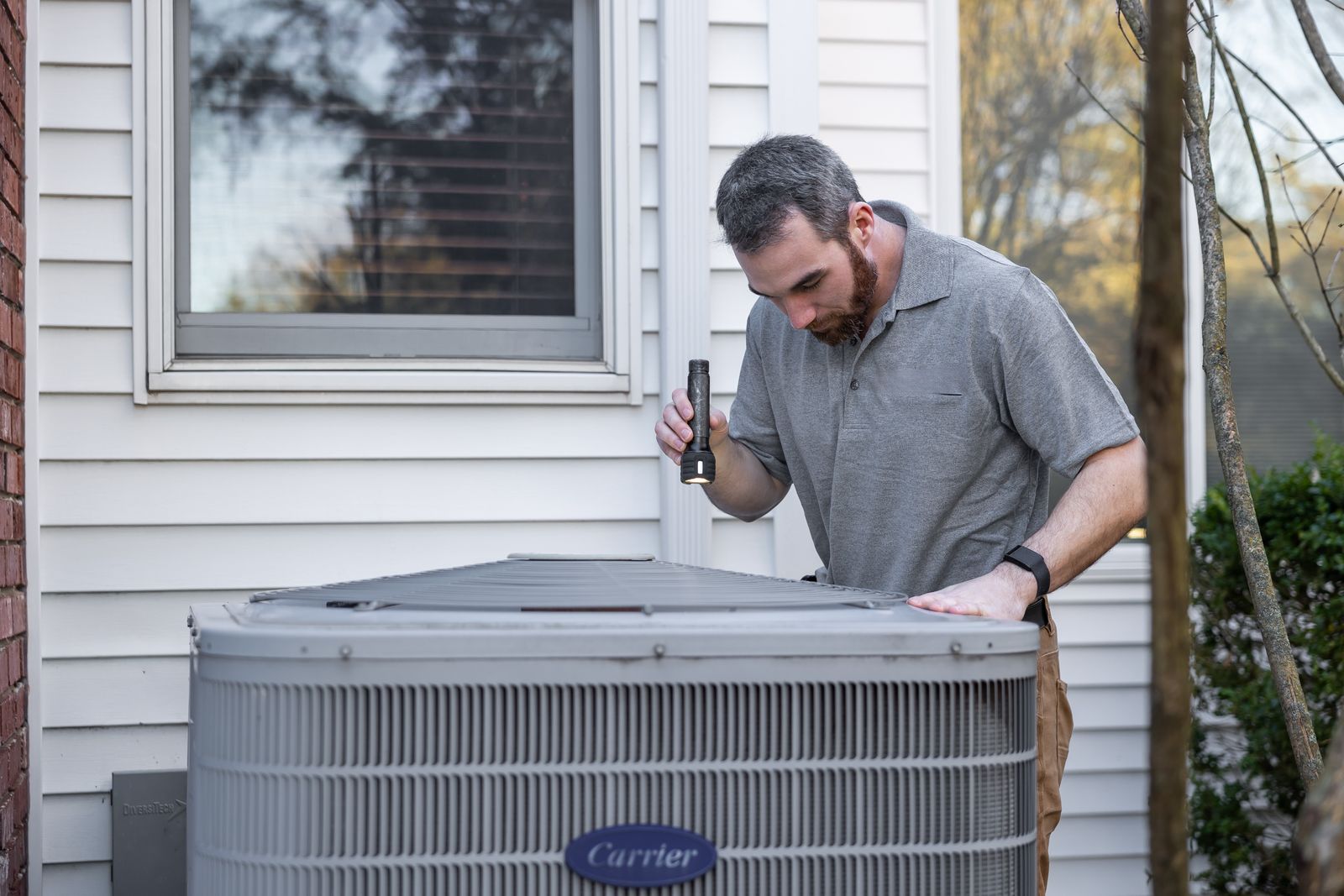 HVAC technician using a flashlight to inspect an outdoor air conditioning condenser unit beside a house