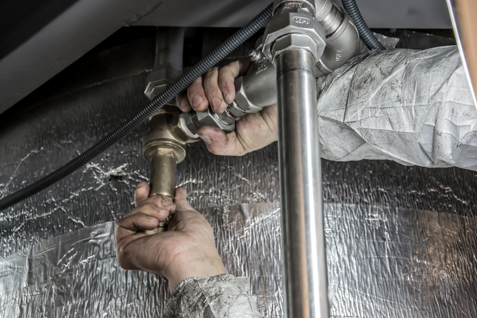 Close-up of a plumber's hands in coveralls tightening a brass pipe fitting under a sink