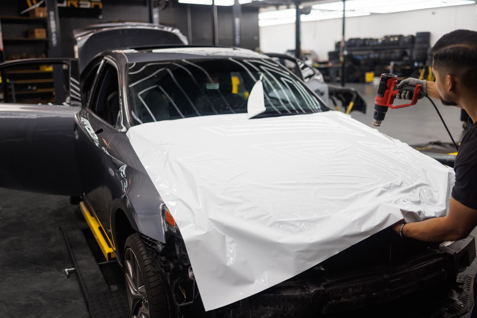 Technician using a heat gun to apply white vinyl wrap film to a car's hood in a shop